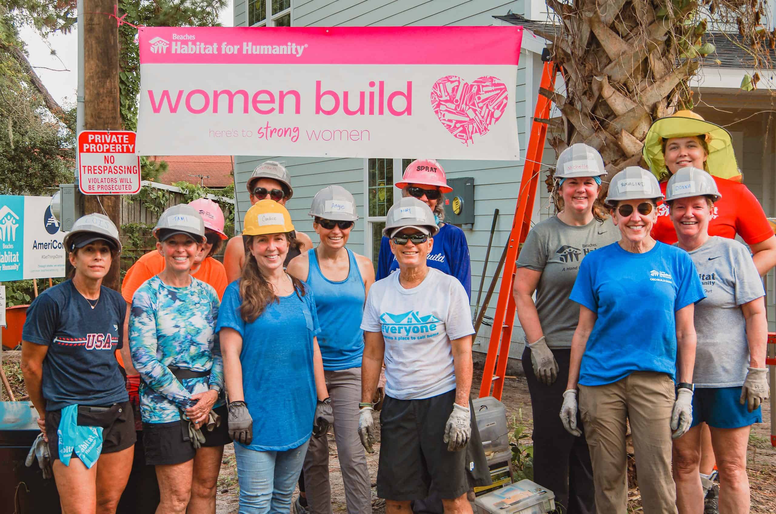 A group of women wearing hard hats and work gloves standing in front of a Beaches Habitat for Humanity ‘Women Build’ banner, smiling together at a build site with a house and construction materials in the background.