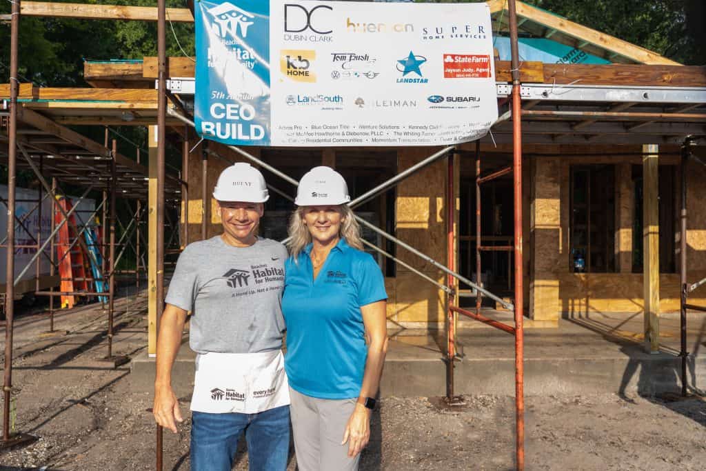Terri Cohen and Brent Paris stand in front of Beaches Habitat's 2024 home on CEO Build day 2024. They are wearing white CEO Build hard hats and standing in front of a CEO Build sign that thanks 2024 CEO Build sponsors.