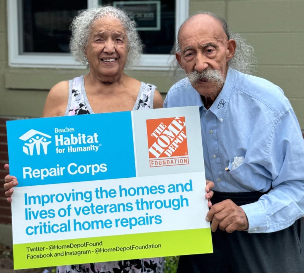 Atlantic Beach couple Cora and Ralph Forney stand in front of their green home with brand new windows. They hold a sign with the Beaches Habitat for Humanity Repair Corps logo, the Home Depot Foundation logo and information about the senior repair program. 